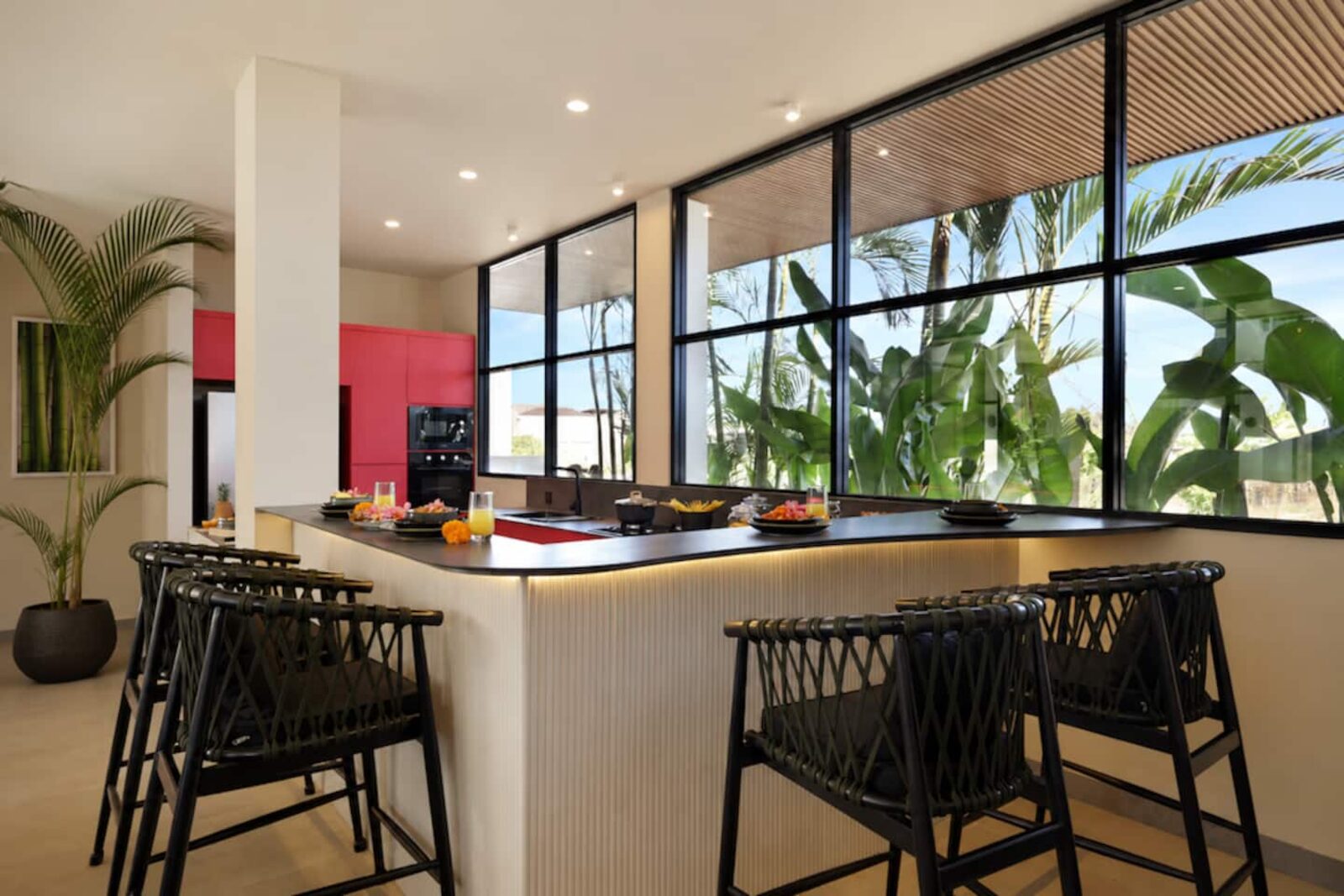 Kitchen island with bar stools under tall windows looking into dense green planting